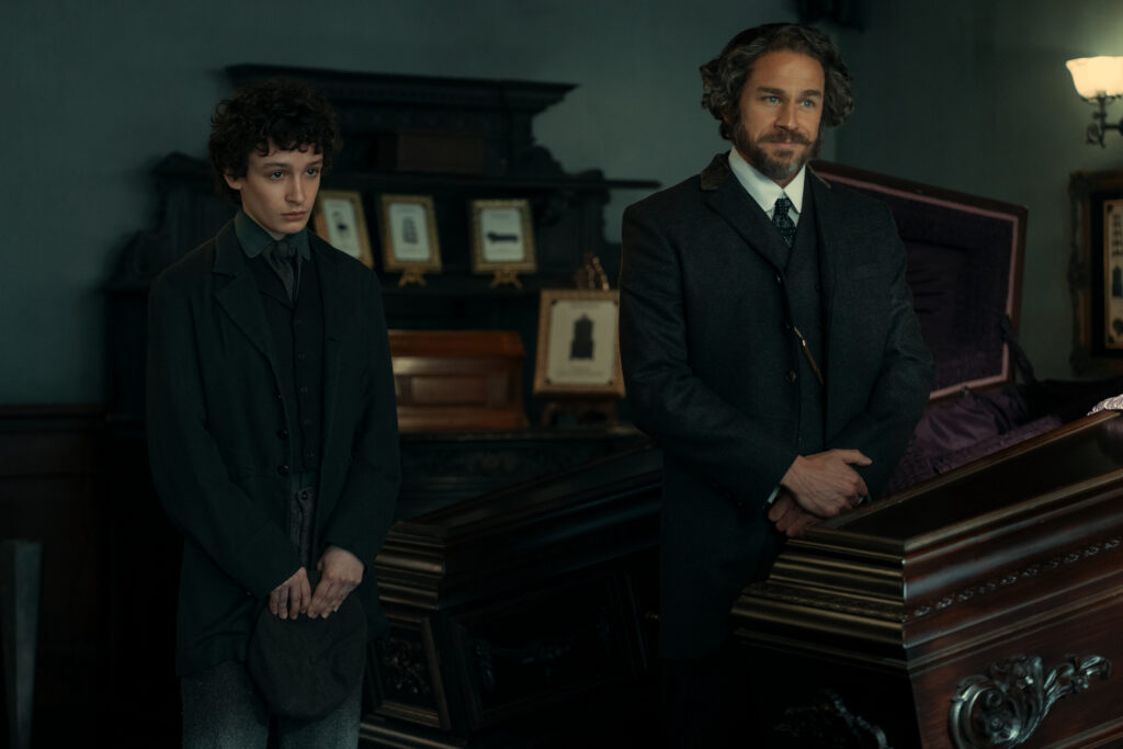 "Two men in dark, old-fashioned suits stand beside a polished wooden coffin in a dimly lit, ornate room decorated with framed photos, suggesting a 19th-century funeral or wake setting."